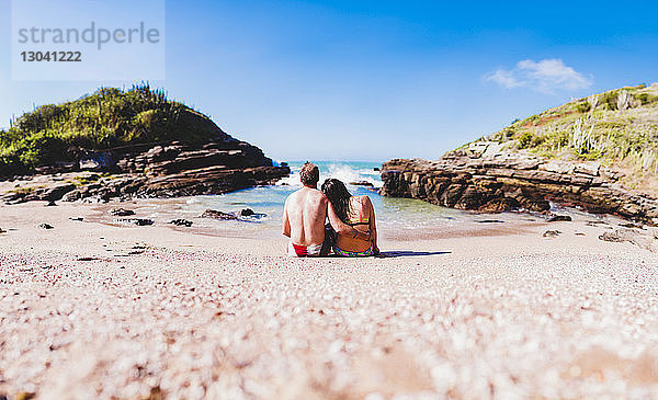 Rückansicht eines anhänglichen Paares  das sich am Strand am Ufer gegen den Himmel entspannt