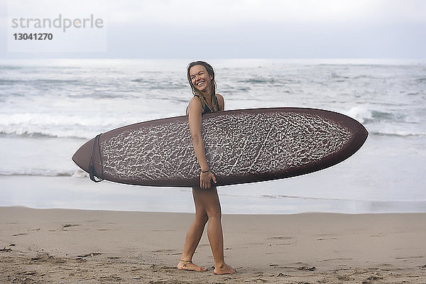 Seitenansicht einer fröhlichen Frau  die beim Strandspaziergang ein Surfbrett trägt