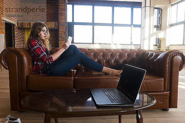 Seitenansicht einer jungen Frau  die einen Tablet-Computer benutzt  während sie zu Hause auf einem Ledersofa sitzt