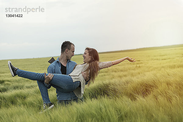 Mann trägt fröhliche Frau auf Grasfeld gegen den Himmel