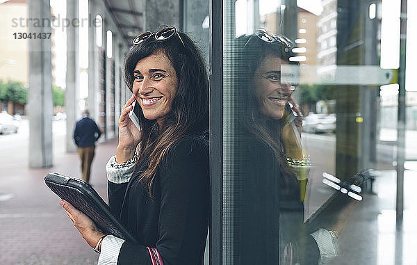 Porträt einer glücklichen Frau  die am Fenster stehend mit dem Handy telefoniert