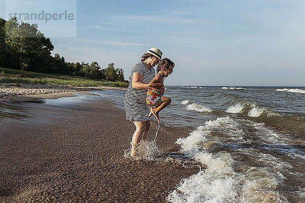 Mutter und Tochter spielen in Wellen am Strand gegen den Himmel
