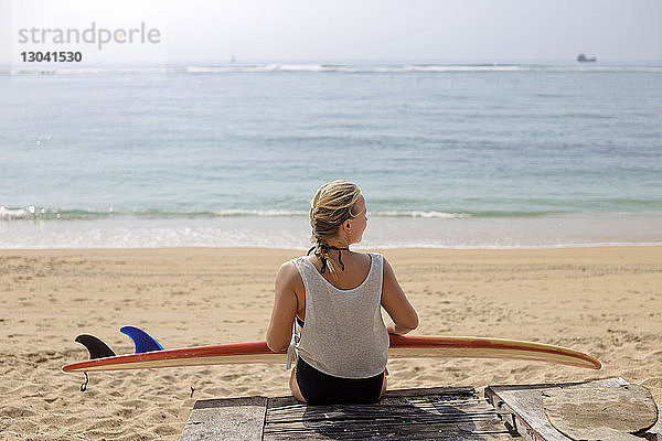 Rückansicht einer am Strand sitzenden Frau mit Surfbrett