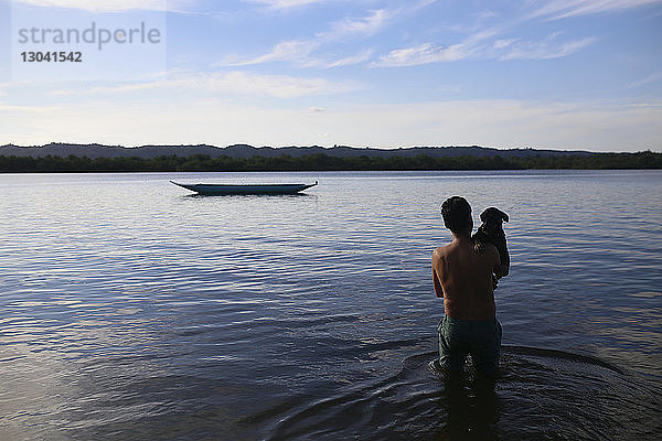 Rückansicht eines hemdlosen Mannes mit Hund  der im See gegen den Himmel steht