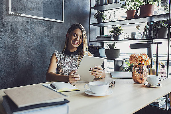 Glückliche Geschäftsfrau benutzt einen Tablet-Computer  während sie im Heimbüro am Tisch sitzt
