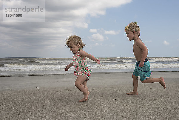 Seitenansicht von verspielten Geschwistern  die am Strand gegen bewölkten Himmel rennen