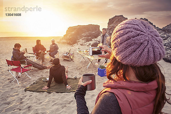 Frau fotografiert Freunde  die bei Sonnenuntergang am Strand zelten