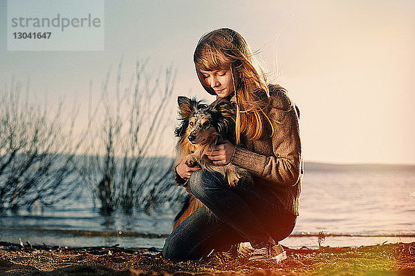 Teenager-Mädchen hält Hund  während sie bei Sonnenuntergang am Strand kniet