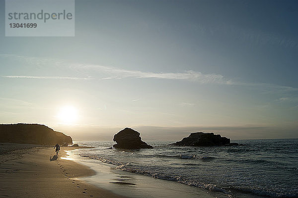 Silhouettenmann in voller Länge mit Surfbrett  der bei Sonnenuntergang am Strand gegen den Himmel läuft