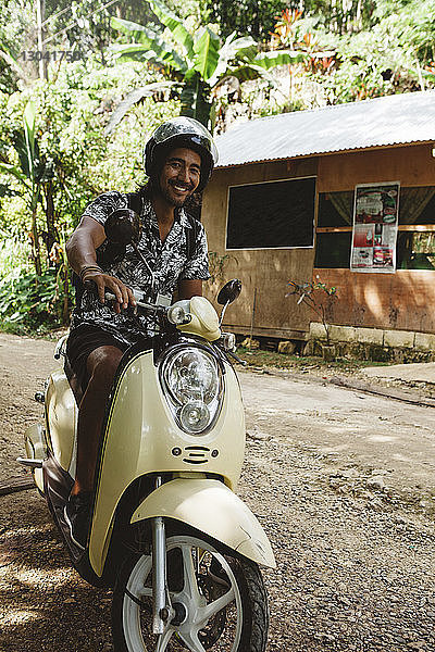 Porträt eines fröhlichen Mannes mit Helm beim Fahren eines Motorrollers auf unbefestigter Straße
