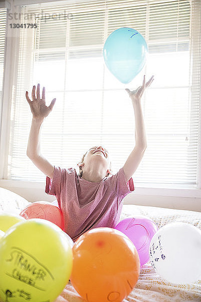 Fröhlicher Junge spielt mit bunten Luftballons  während er zu Hause auf dem Bett sitzt