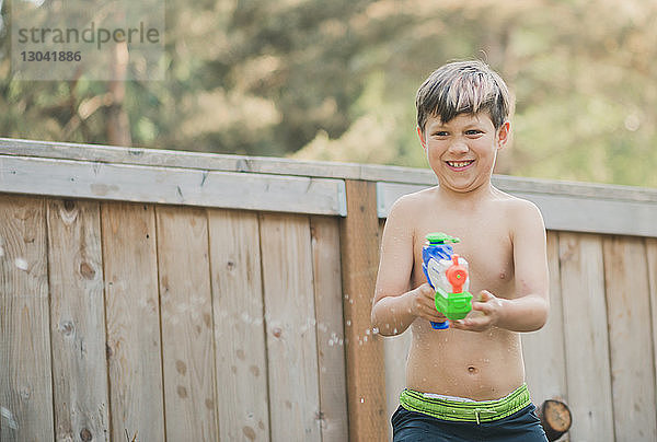 Glücklicher Junge spielt mit Wasserpistole  während er am Holzzaun im Hof steht