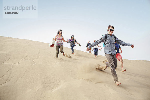Fröhliche Freunde rennen Sanddüne gegen klaren Himmel hinunter