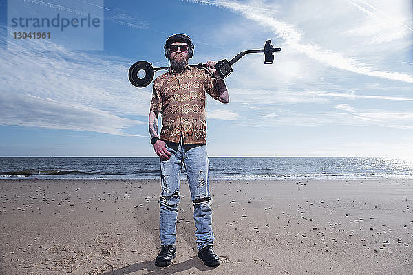 Ganzflächiges Porträt eines Mannes  der den Metalldetektor am Strand von Horsey Beach gegen den Himmel hält