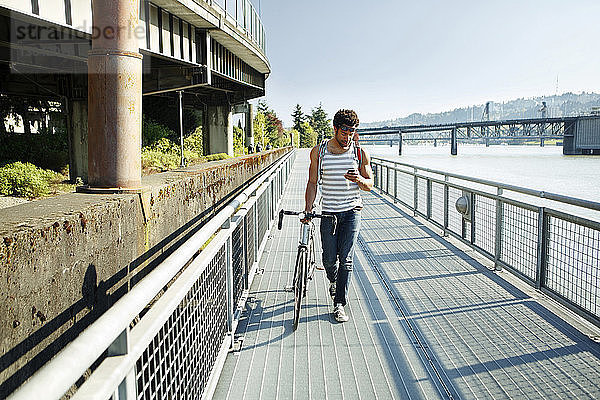 Mann benutzt Mobiltelefon beim Spazierengehen mit Fahrrad auf der Promenade