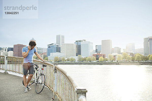 Junger Athlet benutzt Mobiltelefon  während er mit dem Fahrrad auf der Brücke am Fluss steht