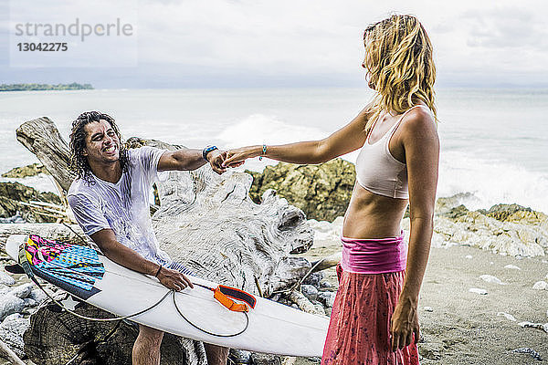 Glücklicher männlicher Surfer hält Frauenhand  während er auf Treibholz am Strand sitzt