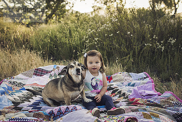 Porträt von Mädchen und Hund auf einer Picknickdecke auf dem Feld sitzend