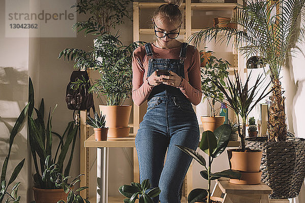 Frau benutzt Smartphone  während sie sich zu Hause an Topfpflanzen an den Tisch lehnt