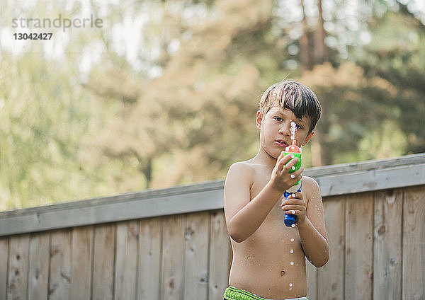 Porträt eines Jungen  der Wasser aus einer Spritzpistole spritzt  während er an einem Holzzaun im Hof steht