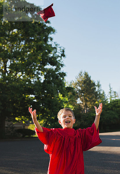 Porträt eines glücklichen Jungen im Abschlusskleid  der Mörtelbrettchen auf den Fußweg wirft
