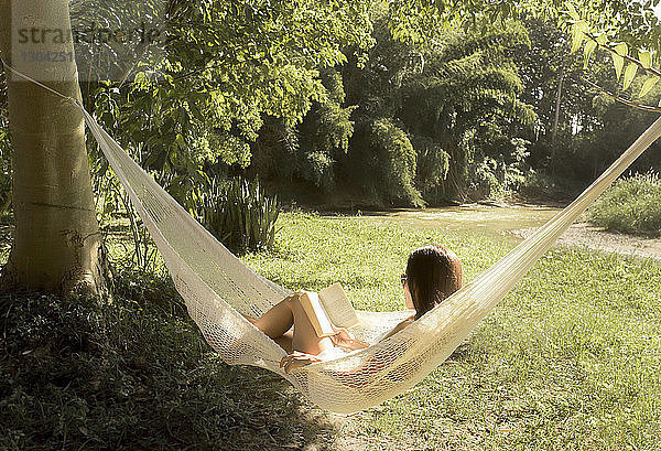 Frau liest ein Buch  während sie sich an einem sonnigen Tag auf einer Hängematte im Park entspannt