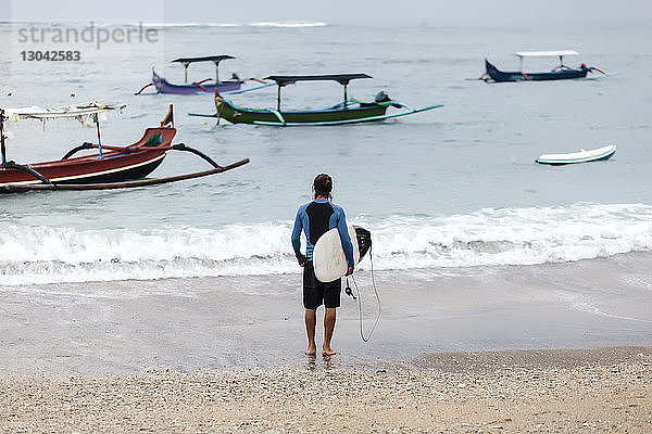 Rückansicht eines Mannes  der aufs Meer schaut  während er ein Surfbrett an Land trägt