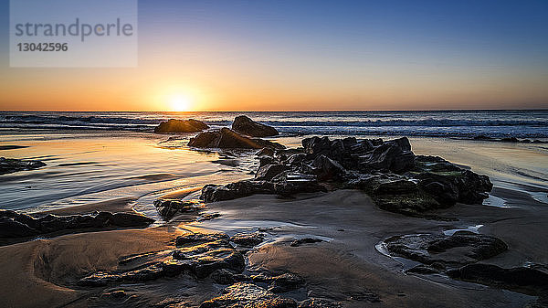 Szenische Ansicht des Meeres mit Felsen am Strand vor dramatischem Himmel bei Sonnenuntergang