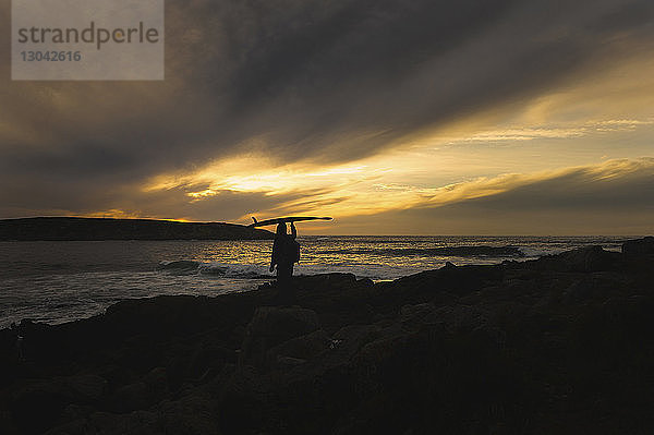 Scherenschnitt eines Mannes  der ein Surfbrett auf dem Kopf trägt  während er bei Sonnenuntergang am Strand steht