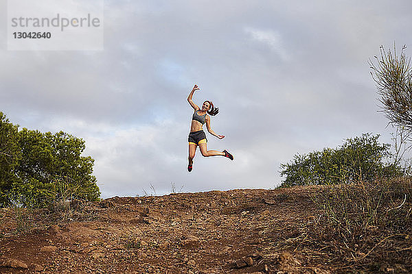 Tiefwinkelansicht einer Frau  die über einen Hügel springt  während sie gegen einen bewölkten Himmel trainiert