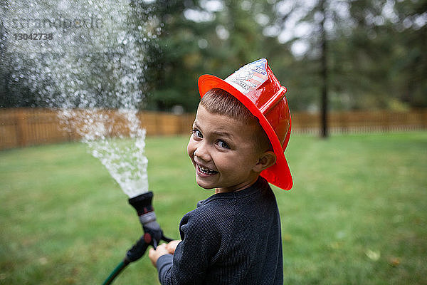 Porträt eines Jungen mit Helm beim Spritzen von Wasser mit dem Gartenschlauch im Hinterhof