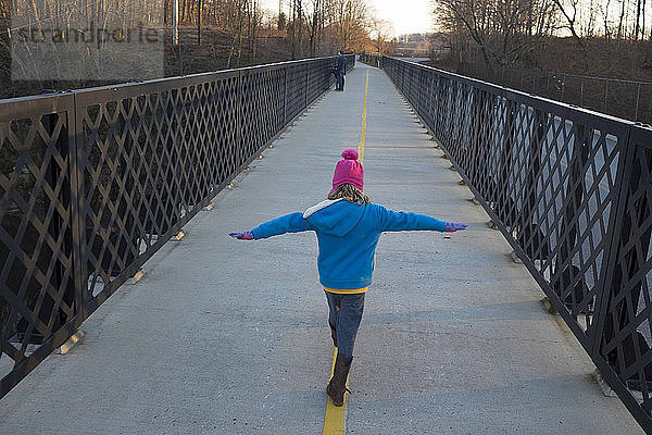 Rückansicht eines verspielten Mädchens mit ausgestreckten Armen beim Gehen auf der Brücke