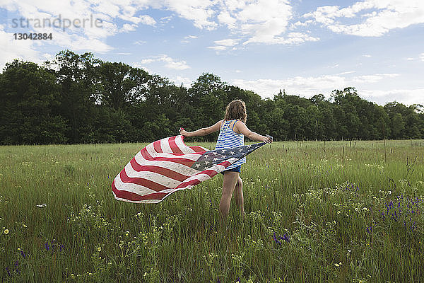 Rückansicht eines Mädchens  das die amerikanische Flagge hält  während es auf einem Grasfeld geht