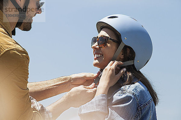 Mann hilft Frau beim Tragen eines Fahrradhelms gegen den klaren Himmel an einem sonnigen Tag