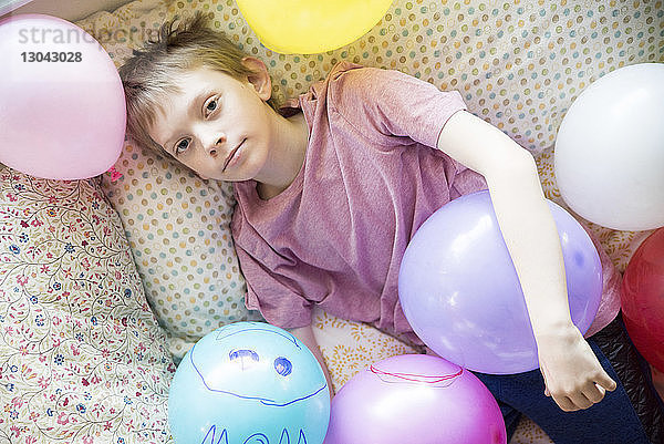 Hochwinkelporträt eines Jungen mit bunten Luftballons  der zu Hause auf dem Bett liegt