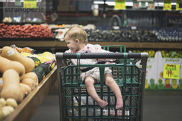 Ein Mädchen in voller Länge sitzt auf einem Einkaufswagen  während es Gemüse vom Marktstand im Supermarkt pflückt