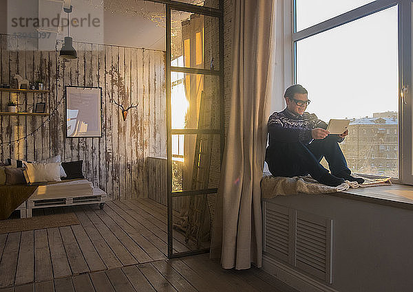Mann benutzt Tablet-Computer  während er zu Hause auf dem Fensterbrett sitzt