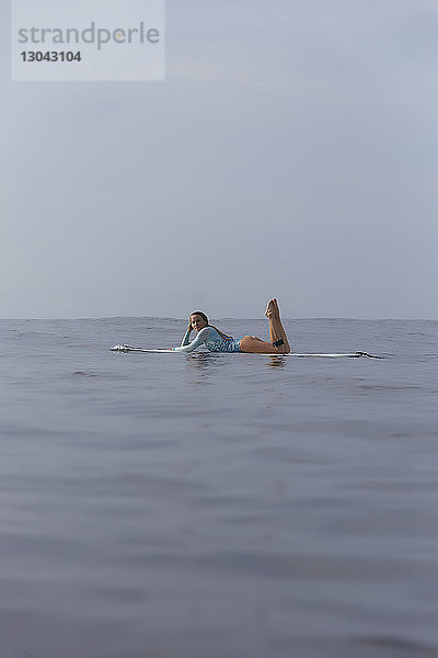 Seitenansicht einer sorgenfreien Frau  die auf einem Surfbrett im Meer gegen den Himmel liegt