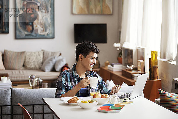 Glücklicher junger Mann benutzt Laptop beim Kaffeetrinken zu Hause
