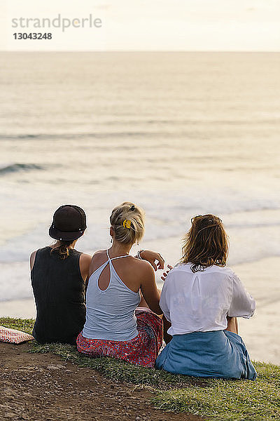 Rückansicht von Freundinnen  die bei Sonnenuntergang am Strand sitzen