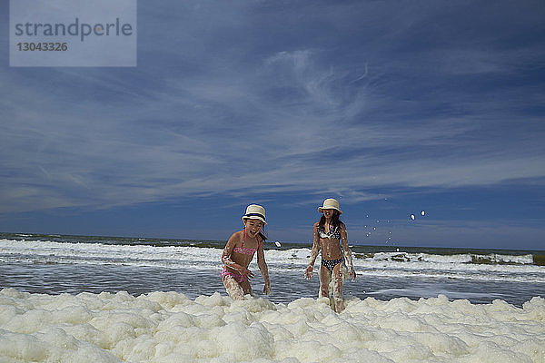 Glückliche Schwestern spielen in Meeresschäumen am Strand gegen den Himmel