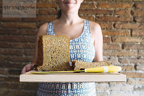 Mittendrin eine Frau  die ein Schneidebrett mit Brot und Küchenmesser gegen eine Ziegelwand hält