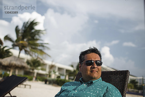 Mann trägt Sonnenbrille  während er sich am Strand auf einem Liegestuhl ausruht