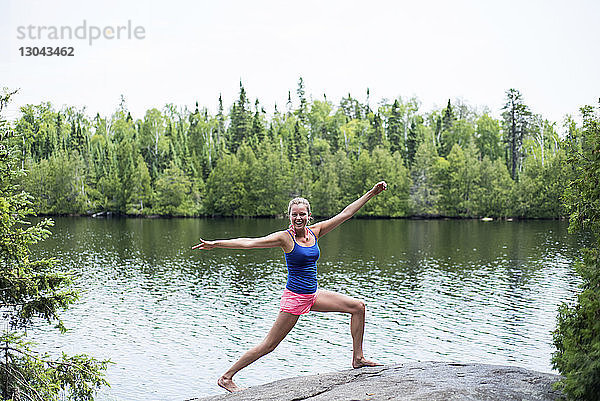 Glückliche Frau praktiziert Yoga  während sie auf einem Felsen am Seeufer vor klarem Himmel steht