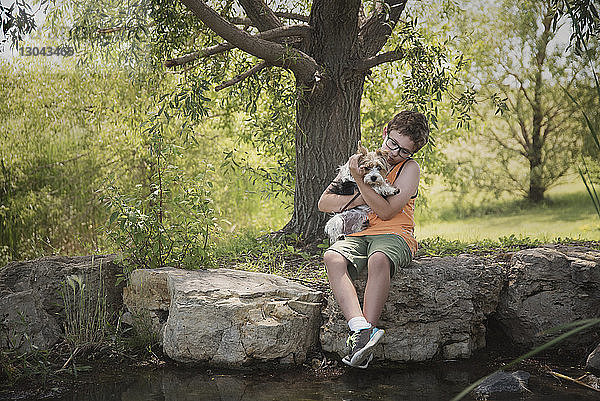 Junge mit Yorkshire Terrier sitzt auf einem Felsen am Teich im Park
