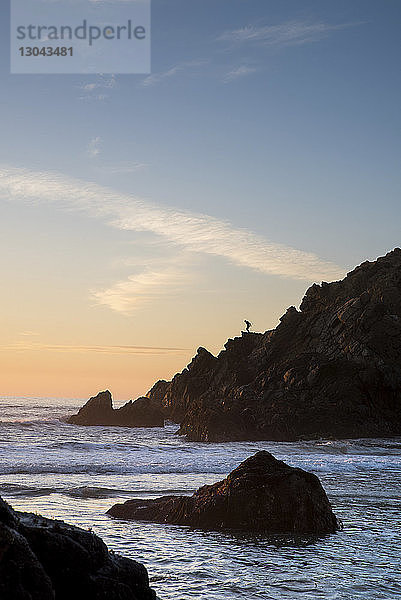 Fernsicht auf die Silhouette eines Teenager-Mädchens  das bei Sonnenuntergang auf einem Berg am Meer gegen den Himmel steht