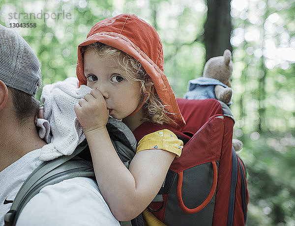 Porträt einer niedlichen Tochter beim Daumenlutschen  während sie vom Vater im Wald getragen wird