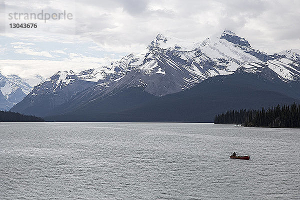 Hochwinkelaufnahme einer Person  die im Boot auf dem Maligne Lake durch schneebedeckte Berge vor bewölktem Himmel im Jasper-Nationalpark segelt
