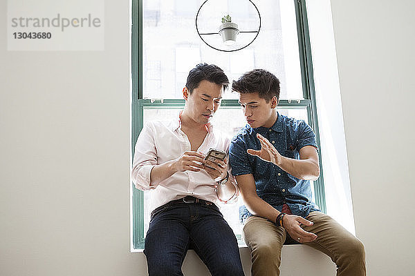 Kreative Geschäftsleute benutzen ein Smartphone  während sie im Büro auf dem Fensterbrett sitzen