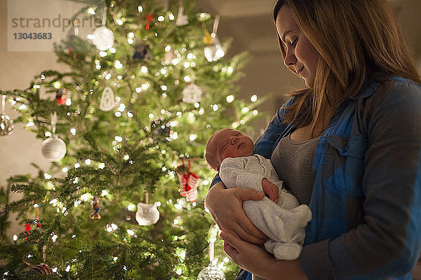 Mutter trägt Neugeborenes  während sie zu Hause am Weihnachtsbaum steht